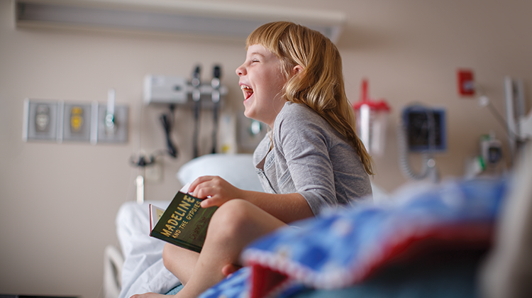 niña hospitalizada riéndose mientras lee un libro en la cama